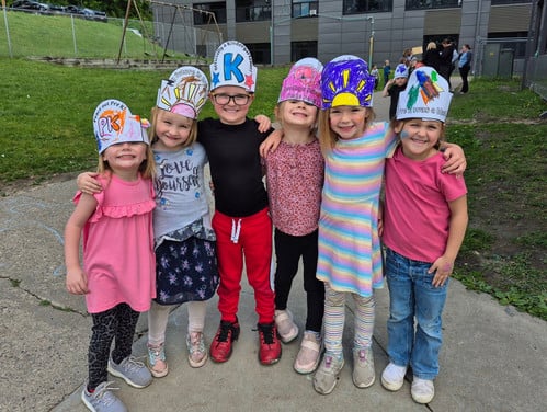 Photo of smiling kindergarten students with their arms around each other wearing paper hats